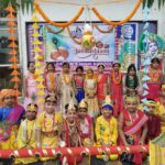 Group of children in bright traditional outfits posing for a festival photo in a schoolyard with marigold garlands and colorful paper decorations behind them